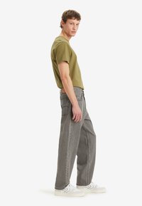 Camiseta verde oliva, vaqueros de pierna ancha grises, zapatillas blancas y grises. El conjunto presenta un diseño casual con un ajuste relajado y una mínima marca.