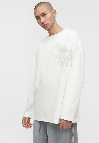 Άνδρας με ξυρισμένο κεφάλι που φοράει ένα λευκό πουκάμισο με μακριά μανίκια με διακριτικό λουλουδάτο σχέδιο στο στήθος και άνετο γκρι παντελόνι.