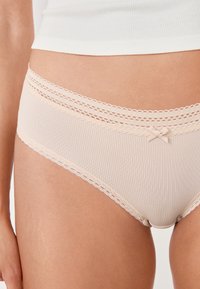 Slip beige a coste con una delicata rifinitura in pizzo e un dettaglio a fiocco in vita, realizzato in un morbido tessuto elasticizzato per un comfort ottimale.
