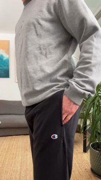 Šedá mikina s volným střihem v kombinaci s černými tepláky, které mají malou modro-červenou značku na kapse levého stehna.