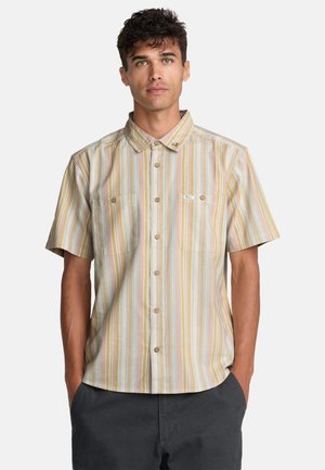 Młody mężczyzna z krótkimi kręconymi włosami, ubrany w pastelową, paskową, krótko rękawową koszulę na guziki i ciemne spodnie, z rękami w kieszeniach, na neutralnym tle.