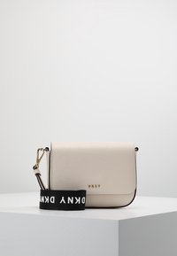 Kreuzkörperlaukku kermaisesta synteettisestä materiaalista, jossa on elävä pintakuvio, musta logovyö, kultaiset metalliosat ja minimalistinen muotoilu.