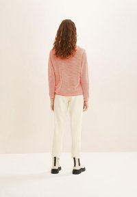 Femme aux cheveux bruns bouclés portant un pull en maille corail, un pantalon blanc et des bottines crème à semelles noires, se tenant devant un fond uni.