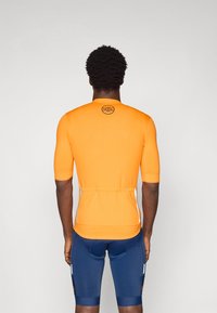 Maillot de cyclisme orange à manches courtes avec un design ajusté, logo au dos et ourlet élastique. Porté avec un short de cyclisme bleu.