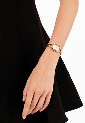 Guldtonet ur med firkantet urskive, hvid skive og kædearmbånd, båret på en hånd mod en sort stofbaggrund.