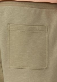 Olivgrüner Stofftaschendetail an der Jogginghose, mit einer horizontalen rechteckigen Form, sichtbaren Nähten und einem kleinen Logoakzent.