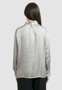 Blusa di raso grigia con collo alto, giromanica rotondo e maniche lunghe. Presenta polsini con bottoni e una texture liscia e lucida.