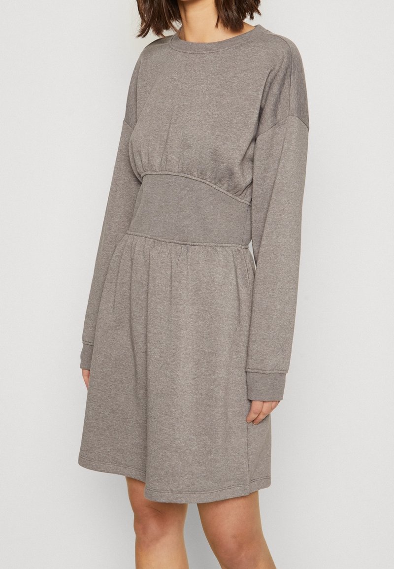 Femme portant une robe décontractée marron clair à manches longues et longueur genou avec un détail froncé à la taille.