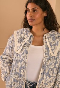 Giacca trapuntata di colore azzurro chiaro con motivo floreale e bordi color crema. Presenta un grande colletto, chiusura con cravatta e un design in tessuto strutturato.