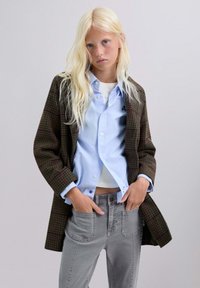 Blond meisje in een geblokte bruine jas, lichtblauwe button-up shirt, witte top eronder en grijze jeans met handen in de zakken.