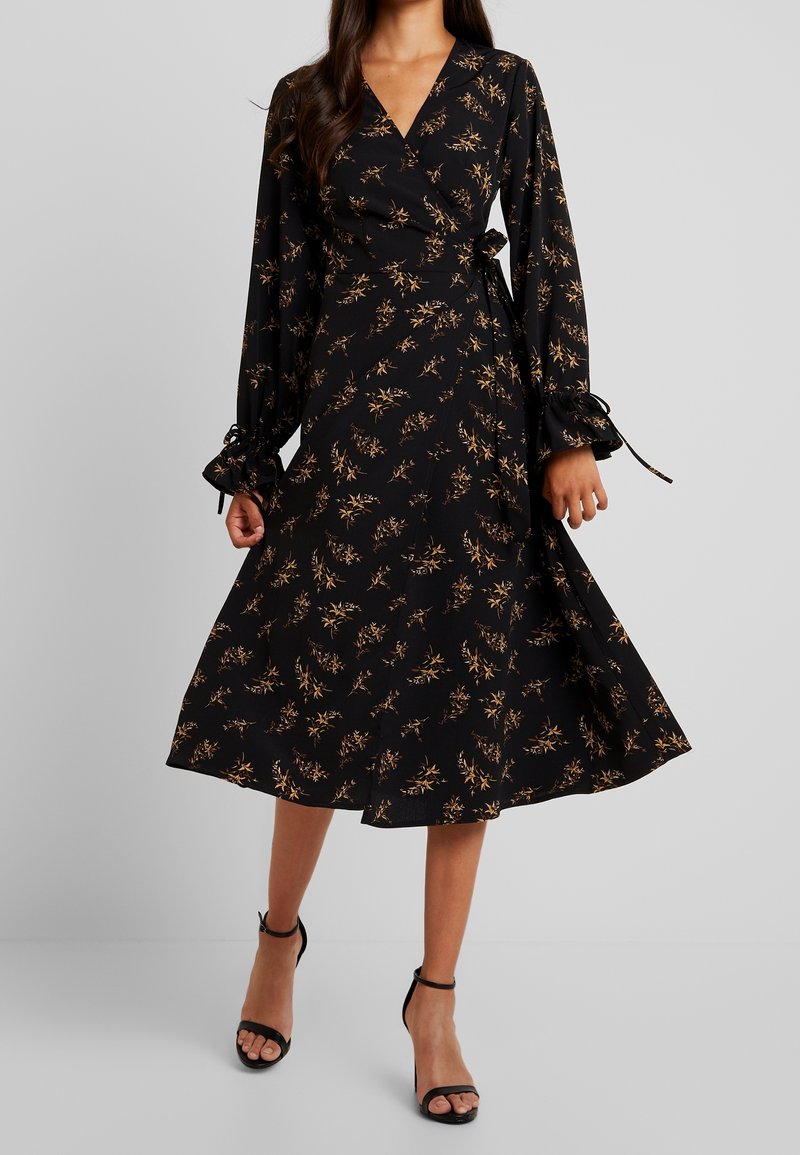 Femme portant une robe portefeuille à manches longues noire à motifs floraux avec des chaussures à talons à bride cheville, debout devant un fond gris uni.