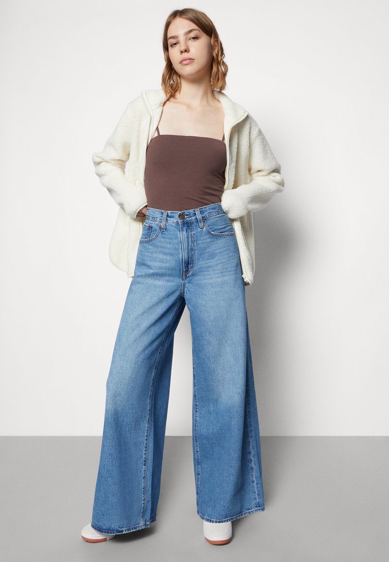 Jean en denim bleu à jambes larges avec une taille haute, associé à un haut ajusté marron et une veste en polaire crème zippée. Chaussures à plateforme blanches.