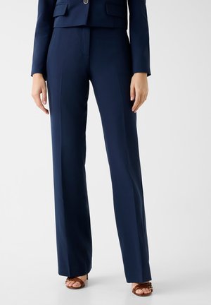 Frau trägt maßgeschneiderte marineblaue Hosen mit weitem Bein und passenden Blazer, kombiniert mit braunen Sandalen mit offenem Zehenbereich und Absatz, stehend vor weißem Hintergrund.