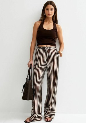 Giovane donna con top nero senza maniche, pantaloni larghi a righe, sandali neri, che porta una borsa intrecciata scura, in piedi contro un muro semplice.