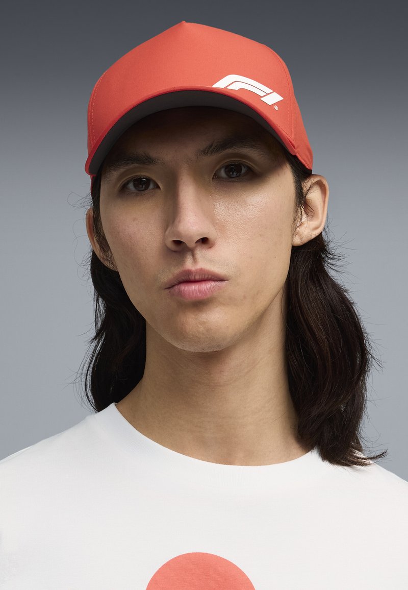 Jeune personne aux longs cheveux foncés porte une casquette rouge avec un logo blanc et une chemise blanche avec un cercle rouge sur la poitrine.