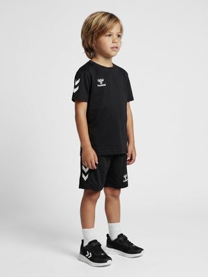 Ungt barn, der står, iført sort sportstøj med hvide chevron-logoer, hvide sokker og sorte sportssko på en ensartet lys baggrund.