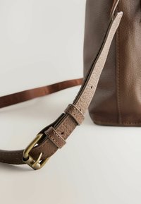 Sangle de sac en cuir marron avec une surface texturée, boucle en métal doré et une accent en cuir. La sangle présente plusieurs trous d'ajustement.