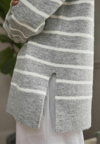 Pull gris avec des rayures blanches, fabriqué en tissu tricot doux. Caractéristiques : fentes latérales et poignets et ourlet côtelés. Détails texturés sur l'ensemble.