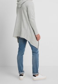 Lysegrå langærmet hættetrøje med buet kant, kombineret med blå cowboybukser og hvide sneakers med sort accent ved hælen.