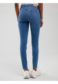 Ļoti piegulošas gaiši zilas džinsa bikses ar kabatām aizmugurē, ko valkā stāvoša persona, kopā ar baltiem biezpědiem sporta apaviem, vienkrāsainā fonā.