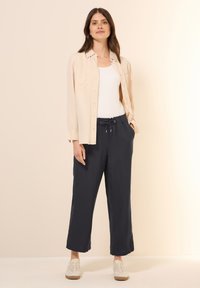 Beige overhemd met knopen over een witte tanktop, gecombineerd met donkergrijze, losse joggingbroeken met katoen en lichte sneakers.