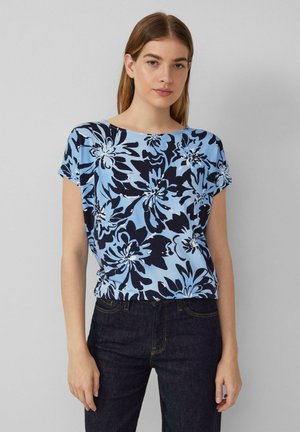 Bloemtop in lichtblauw met donkerblauwe en witte florale patronen, korte mouwen, ronde halslijn en een ontspannen pasvorm, gecombineerd met donkere jeans.