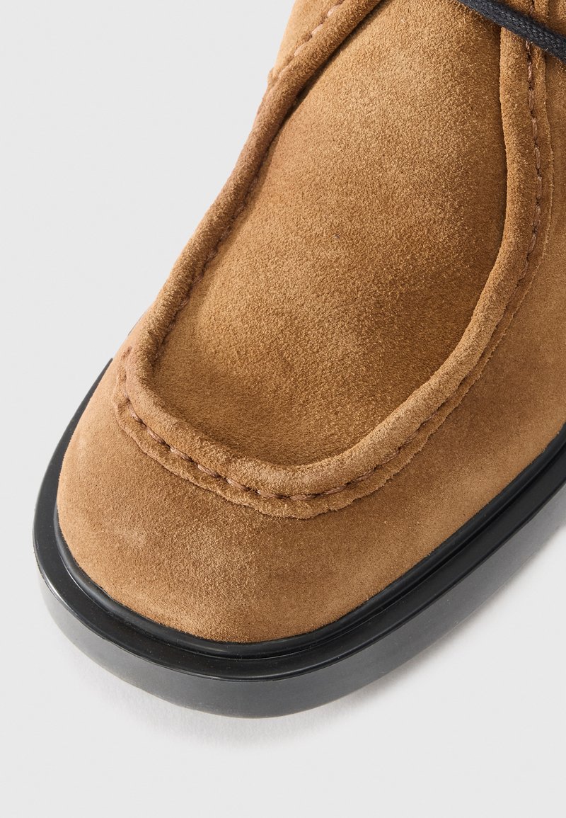 Braunes Wildlederschuh mit abgerundetem Zeh, ausgestattet mit einer schwarzen Gummisohle und sichtbaren Nähten entlang der Kante. Glatte Textur mit minimalen Akzenten.