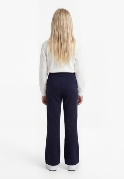 Niño con cabello largo y rubio está de espaldas, vistiendo una camiseta blanca de manga larga, pantalones azul marino y zapatos blancos sobre un fondo blanco.