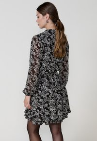 Rochie neagră cu flori, cu mâneci lungi dintr-un material transparent, tiv ondulat și talie strânsă. Are un model floral delicat alb pe fundal.