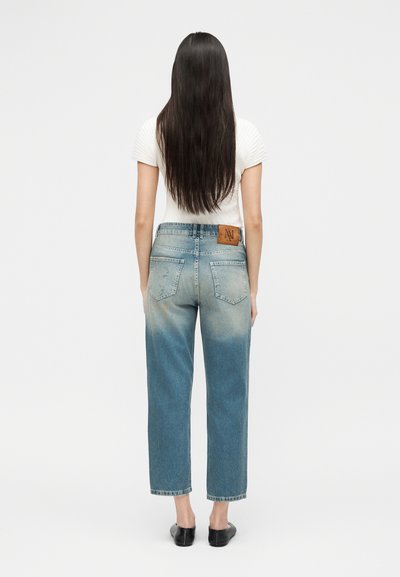Femme aux longs cheveux bruns portant un haut blanc à manches courtes, un jean bleu délavé taille haute et des chaussures noires sans lacets, debout de dos.