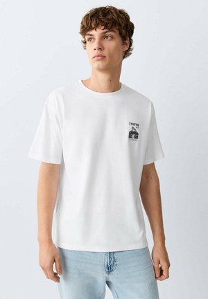 Jonge man met krullend haar die een wit T-shirt draagt met een kleine Tokyo-afbeelding op de borst en lichtblauwe spijkerbroek tegen een effen achtergrond.