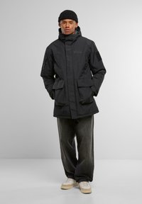 Černá izolovaná bunda s kapucí, několika předními kapsami a zapínací boční kapsou. V kombinaci s širokými tmavými džínami a bílými teniskami.