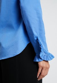 Right arm in blue blouse with ruffled cuff resting next to black pants on light background.