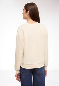 Femme portant un sweat-shirt crème et un jean bleu, vue de dos sur un fond blanc uni.