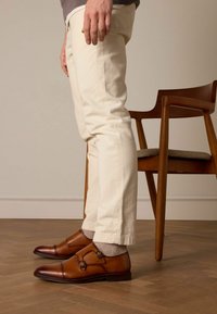 Chaussures en cuir marron à double boucle avec des bouts ronds. Portées avec un pantalon de couleur claire et accentuées par des chaussettes grises claires, sur un sol en bois.