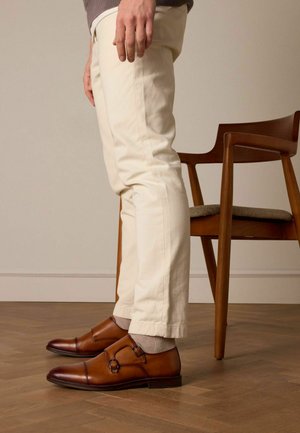Chaussures en cuir marron à double boucle avec des bouts ronds. Portées avec un pantalon de couleur claire et accentuées par des chaussettes grises claires, sur un sol en bois.