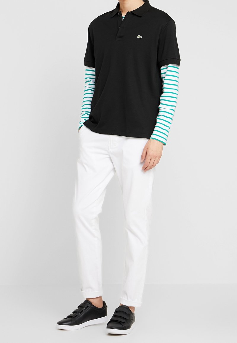Personne portant un polo noir sur une chemise blanche à rayures vertes à manches longues, un pantalon blanc et des baskets noires à semelles blanches.