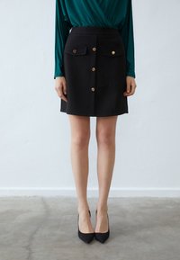 Černá sukně ke kolenům vyrobená z hladké látky, vybavená dvěma předními kapsami, zlatými detailem knoflíků a přiléhavým střihem. V kombinaci s černými vysokými podpatky.