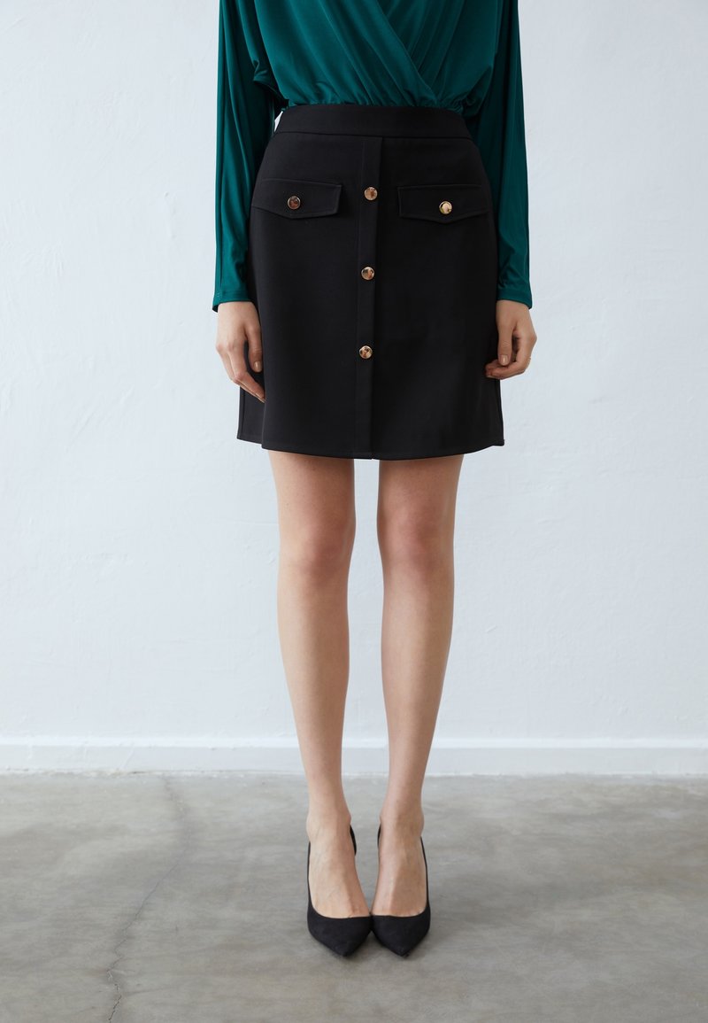 Černá sukně ke kolenům vyrobená z hladké látky, vybavená dvěma předními kapsami, zlatými detailem knoflíků a přiléhavým střihem. V kombinaci s černými vysokými podpatky.