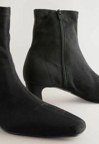 Bottines en daim noir avec un bout pointu et un talon bas. Elles comportent une fermeture éclair sur le côté et des détails cousus sur le dessus.