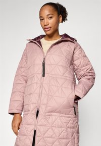 Manteau long matelassé rose clair avec zip, capuche et poches latérales. Motif en losanges texturé et doublure intérieure sombre contrastante.