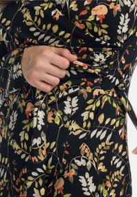 Robe portefeuille fleurie avec une base noire, ornée d'un motif de feuilles orange, vertes et crème, fabriquée dans un tissu doux et extensible.