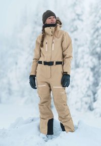 Tuta da neve impermeabile beige con cintura, tasche con zip e dettagli neri. Indossata con guanti grigi e un cappello di lana marrone, ambientata in un paesaggio innevato.
