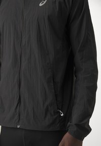 Veste légère noire avec une fermeture éclair frontale, des poches latérales, des poignets élastiques et un petit logo Asics près de l'épaule gauche.