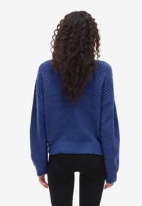 Tekstūruotas mėlynas megztinis su plačiomis, raukšlėtomis pečių linijomis ir ilgomis rankovėmis, pasižymintis laisvu siluetu. Dėvimas su juodais kelnais, matomas iš nugaros.