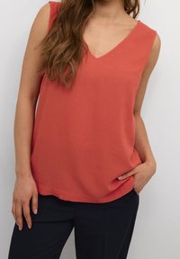 Coral sleeveless top with a V-neck design, made of soft fabric. Features a relaxed fit and a straight hemline, paired with black pants.