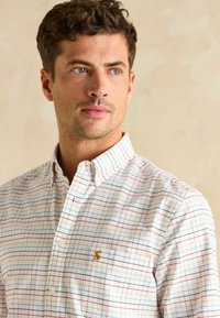 Košeľa s gombíkmi a károvaným vzorom v červenej, modrej a bielej farbe. Obsahuje zlaté logo na vrecku na prsiach a stojatý golier.