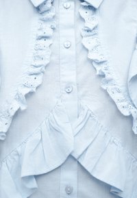 Γαλάζιο πουκάμισο με κουμπιά, με φλοράλ δαντελένια βολάν κατά μήκος της σειράς των κουμπιών και υφασμάτινα βολάν που διασχίζουν το μέσο.