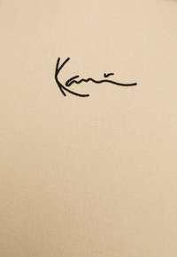 Beige bomullstyg med en slät textur som pryds av en svart broderad signatur "Kani" i kursiv stil.