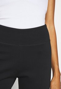 Černé legíny se širokým pasem, hladkým povrchem a zesíleným šitím. Na vršku je bílá přiléhavá košile.